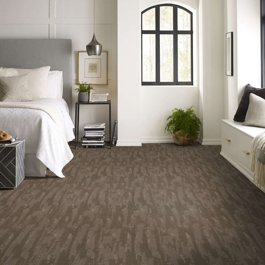 A modern bedroom with neutral tones, featuring a gray bed, white bedding, a small black side table with decor, a large window, a green plant, and textured brown carpet flooring.