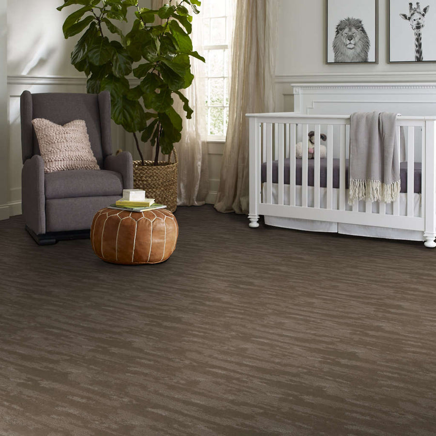 A cozy nursery with a crib, a gray armchair with a pink pillow, a round brown leather ottoman, a large leafy plant, and animal art on the wall, all set on a brown carpeted floor.