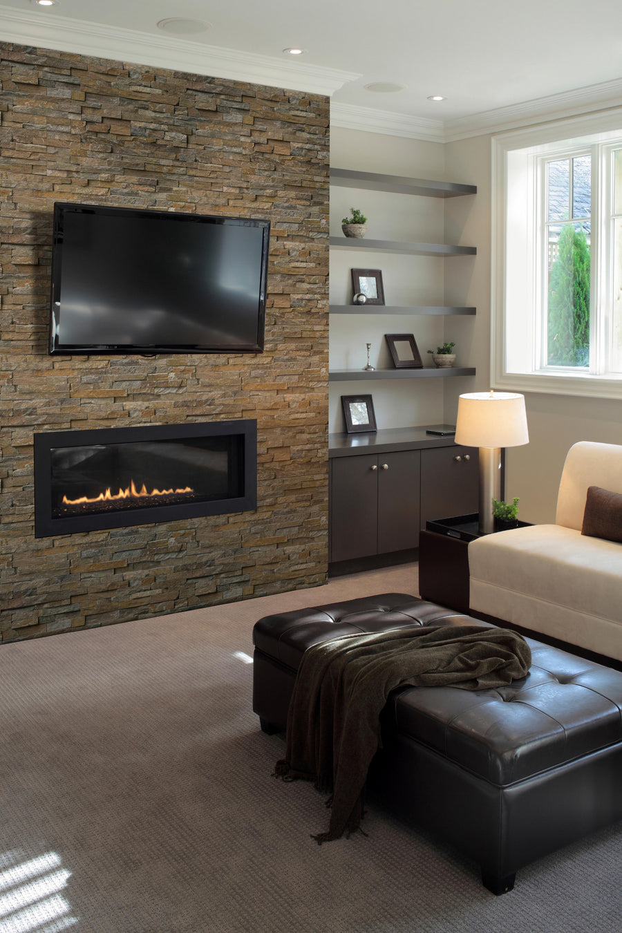 Modern living room with a stone accent wall featuring a mounted flat-screen TV and rectangular fireplace, cream sofa, dark leather ottoman, built-in shelves, and large window letting in natural light.