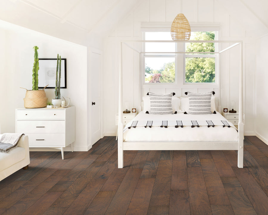 A bright, airy bedroom with white walls and ceiling, a white four-poster bed with striped pillows, a white dresser with a potted cactus, wooden floors, and large windows letting in natural light.