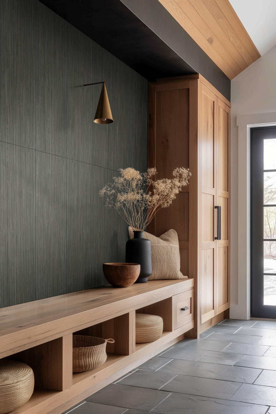 A modern entryway with wooden cabinets, a built-in bench, woven baskets, and a vase with dried foliage on a gray tiled floor. A black wall sconce and natural light from a glass door complete the scene.