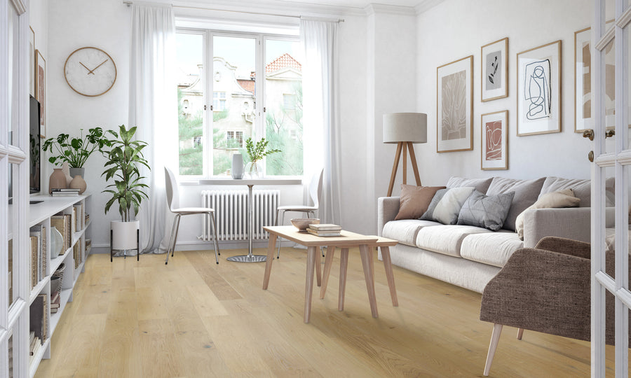 Bright, modern living room with light wood floors, white walls, a beige sofa, brown armchair, wooden coffee table, floor lamp, plants, and framed artwork. Large window lets in natural light.