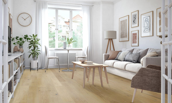Bright, modern living room with light wood floors, white walls, a beige sofa, brown armchair, wooden coffee table, floor lamp, plants, and framed artwork. Large window lets in natural light.
