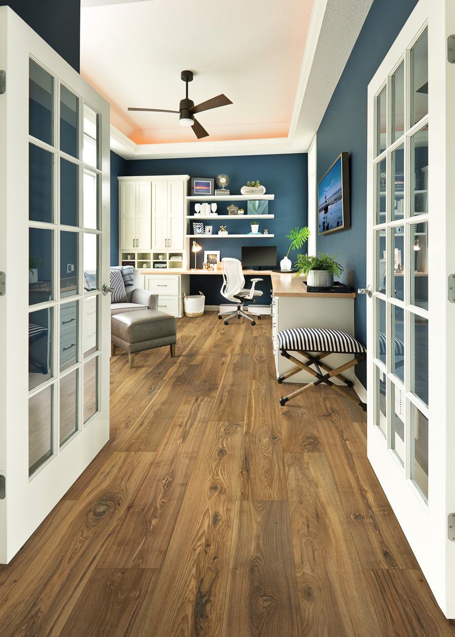 A modern home office with blue walls, wood flooring, French doors, built-in shelves, a desk with a computer, a white office chair, and a striped ottoman, with plenty of natural light.
