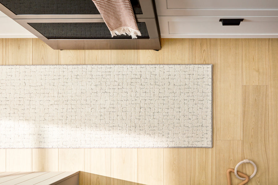 A top-down view of a light-colored woven rug on a wooden floor next to white cabinets. Part of a striped towel hangs from a drawer, and a wooden ring toy is on the floor in the bottom right corner.