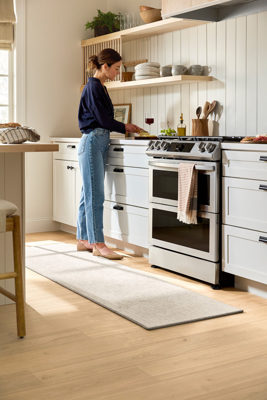 A woman stands in a bright, modern kitchen preparing food at the stove. Open shelves display dishes, a glass of red wine sits nearby, and sunlight streams through a window, illuminating the space.