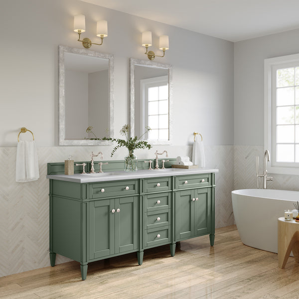 A modern bathroom with light green double-sink vanity, two mirrors, wall sconces, a freestanding white bathtub, herringbone tile accents, and a wooden floor. A vase with greenery sits on the vanity.