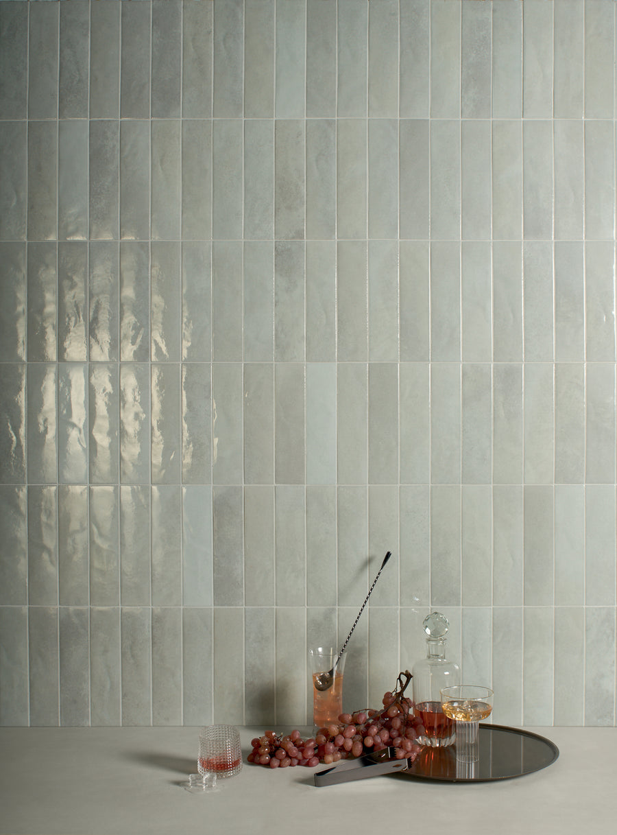 A minimalist scene with a light grey tiled wall, a low table holding a glass of red liquid, a cocktail, a glass decanter, two glasses of white wine, a bunch of red grapes, and a round metal tray.