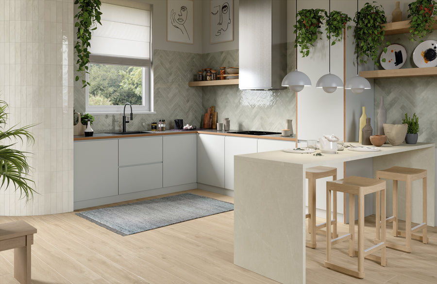 Modern kitchen with light wood flooring, white cabinets, a large island with wooden stools, hanging pendant lights, greenery, and a window with a garden view. Decorative plates and artwork add stylish touches.