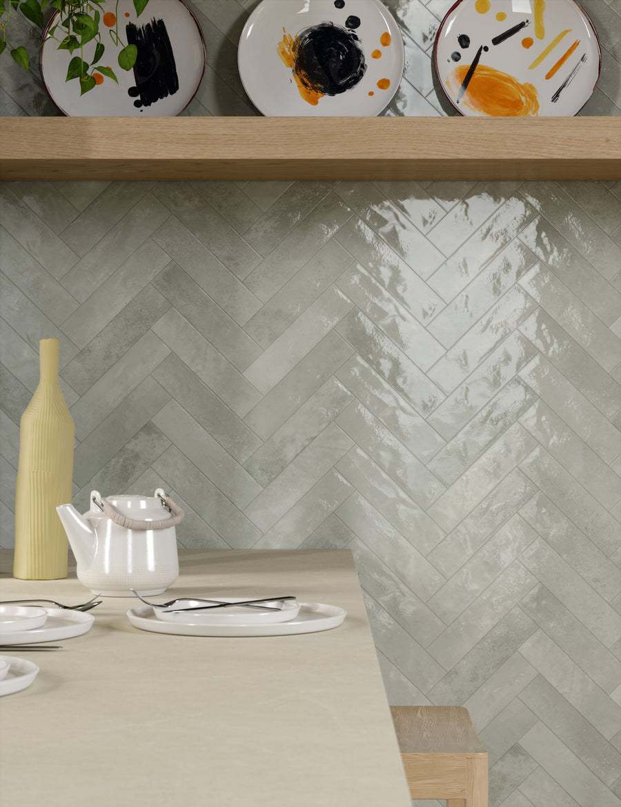 A modern kitchen with a glossy, light gray herringbone tile backsplash, a floating wooden shelf holding artistic plates, and a countertop set with a teapot, plates, cutlery, and a yellow vase.