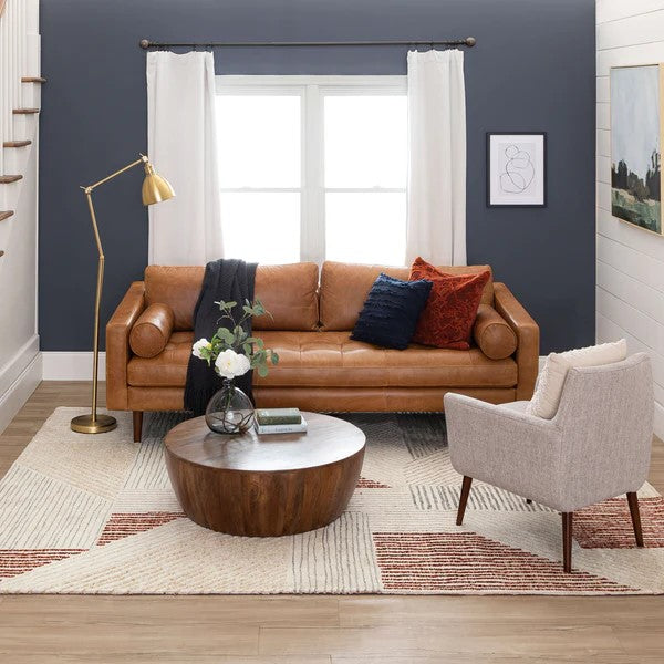 A modern living room with a tan leather sofa, a round wooden coffee table, a light gray armchair, and a gold floor lamp. The room has a geometric rug, dark gray accent wall, and white curtains over a window.
