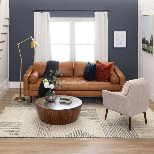 A modern living room with a tan leather sofa, a gray armchair, and a round wooden coffee table on a geometric rug. The sofa has pillows and a throw, and a gold floor lamp stands beside it. White curtains frame a window.