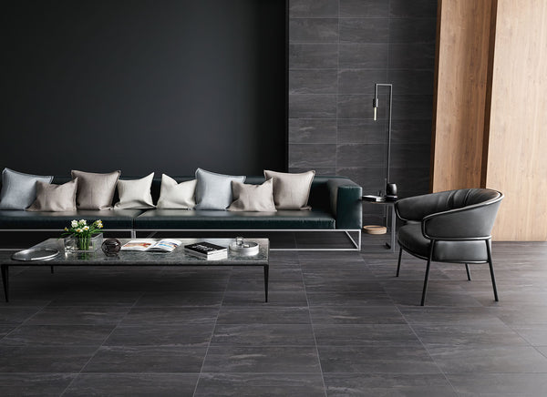 Modern living room with a long dark green sofa decorated with neutral pillows, a black armchair, a sleek black coffee table with magazines and flowers, and dark gray stone-look tile flooring and walls.