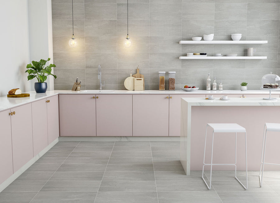 Modern kitchen with light pink cabinets, gray tile walls and floors, white countertops, two pendant lights, open shelves with dishes, a potted plant, and two white bar stools at the island.
