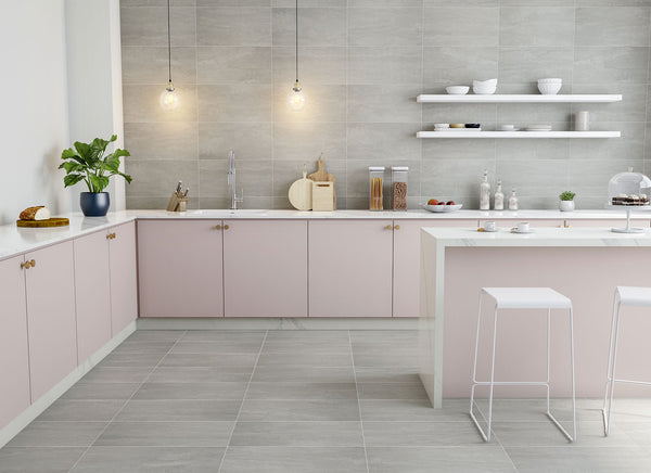 Modern kitchen with light pink cabinets, gray tile walls and floors, white countertops, two pendant lights, open shelves with dishes, a potted plant, and two white bar stools at the island.