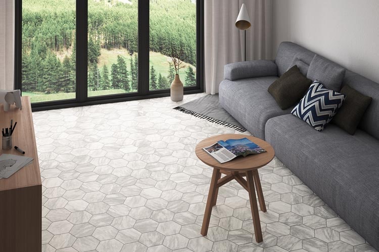 A modern living room with a gray sofa, geometric cushions, a round wooden coffee table, and hexagonal tile flooring. Large windows reveal a view of green pine trees outside.