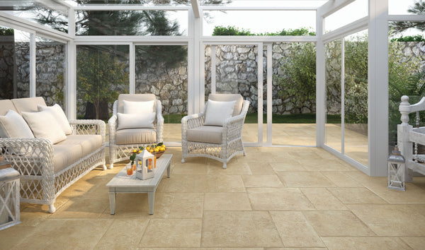 A bright sunroom with wicker furniture, including a sofa and armchairs with cream cushions, a glass coffee table with candles and drinks, and large windows overlooking a stone wall and garden.