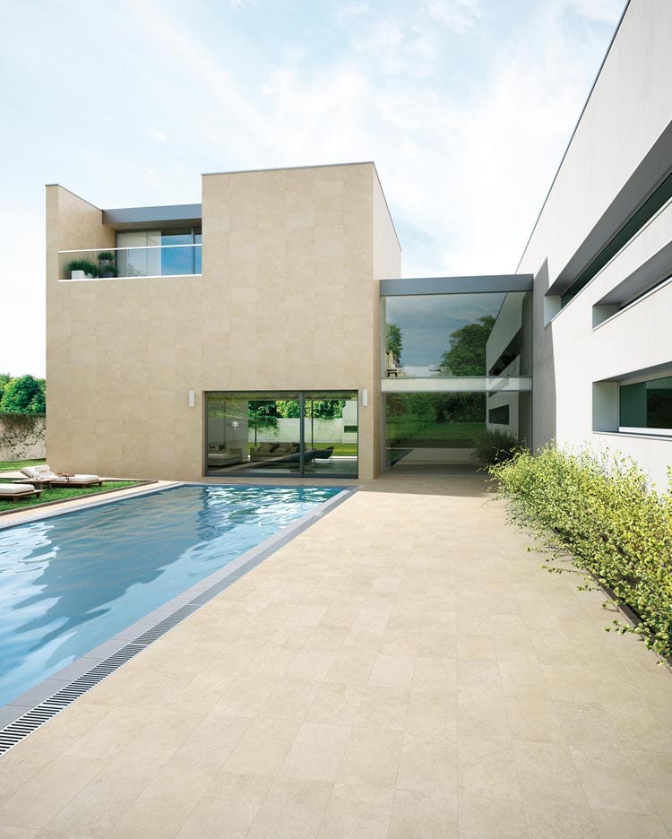 Modern beige and white house with large windows, adjacent to a long rectangular swimming pool, surrounded by greenery and a spacious tiled patio under a partly cloudy sky.