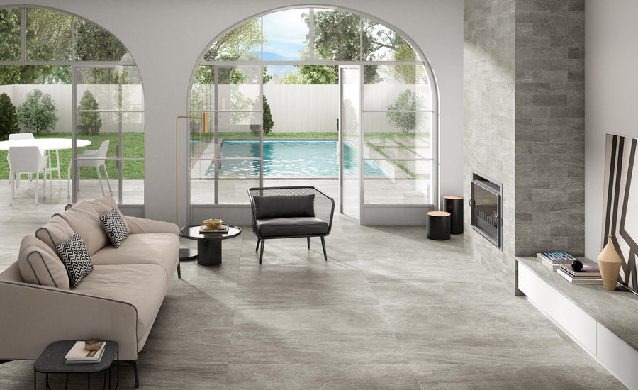 A modern living room with a beige sofa, black armchair, fireplace, and large arched windows overlooking a backyard with a pool, patio, and green lawn. Light streams in through the open glass doors.