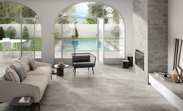 A modern living room with a beige sofa, black armchair, fireplace, and large arched windows overlooking a backyard with a pool, patio, and green lawn. Light streams in through the open glass doors.