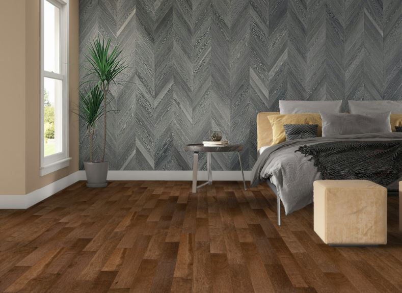 Modern bedroom with a herringbone-patterned gray accent wall, wooden floor, a bed with gray and yellow bedding, a small bedside table, potted plant by the window, and a beige ottoman.