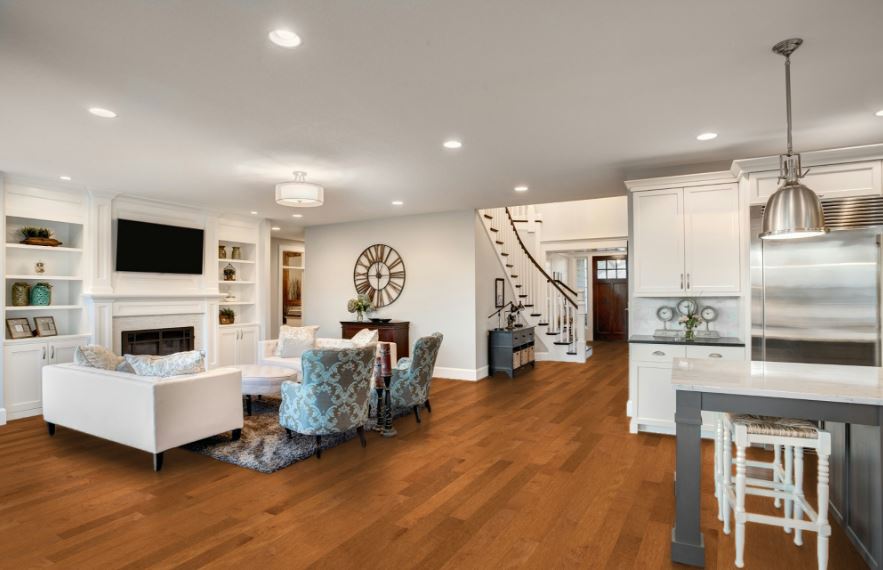 Open-concept living room and kitchen with hardwood floors, white cabinets, stainless steel appliances, a fireplace, a large wall clock, and a seating area with sofas and armchairs. Stairs lead to an upper floor.