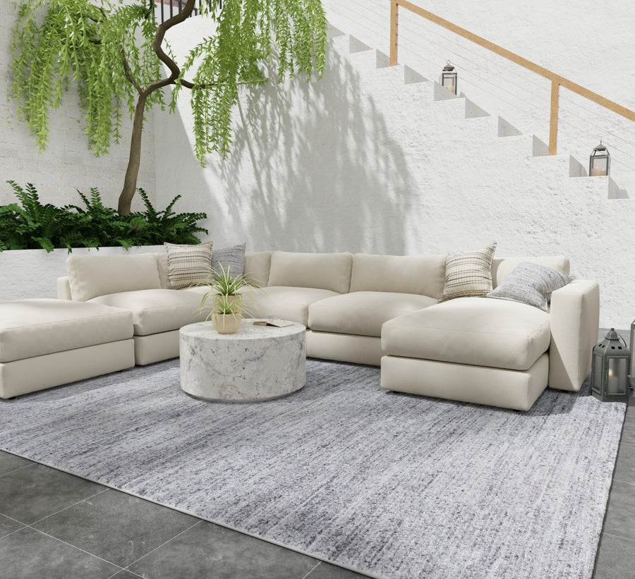 Outdoor seating area with a large cream sectional sofa, a round marble coffee table, and a light gray rug on dark tile flooring. There are green plants and a white textured wall with stairs and a wooden handrail in the background.