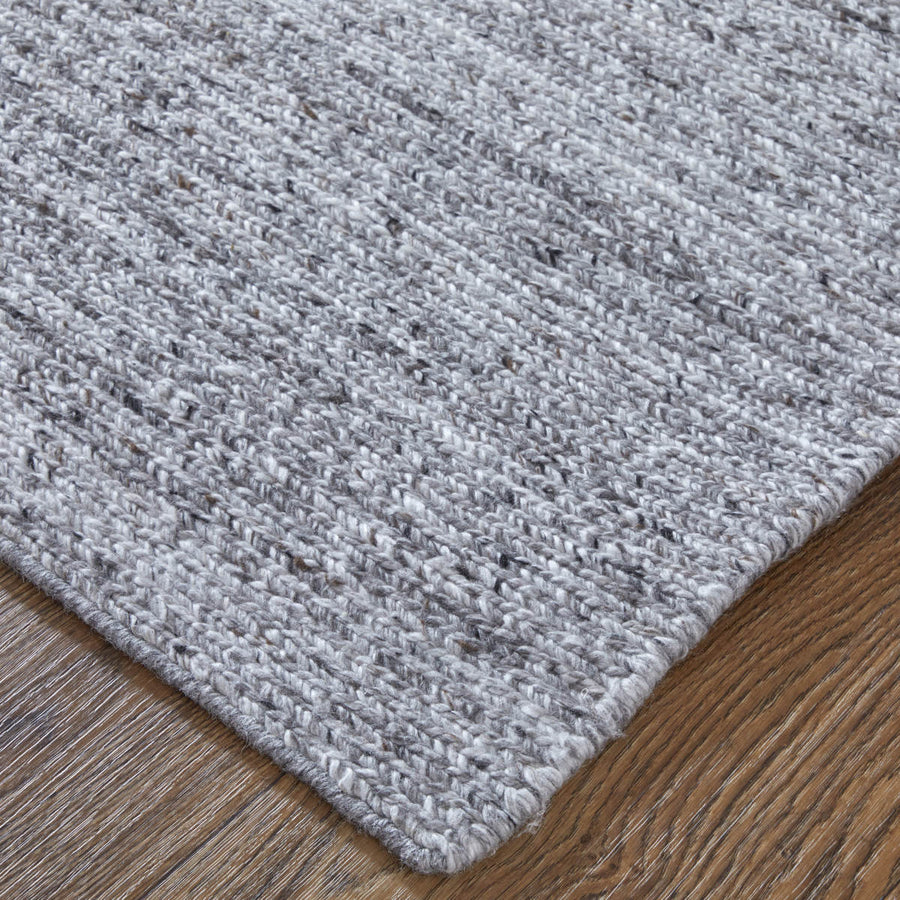 A close-up of the corner of a textured light gray woven rug placed on a wooden floor, showing the rug’s detailed fibers and subtle color variations.