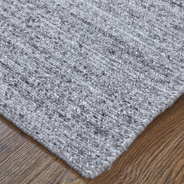 A close-up of the corner of a textured light gray woven rug placed on a wooden floor, showing the rug’s detailed fibers and subtle color variations.