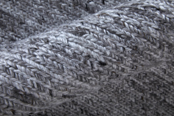 Close-up of a soft, gray knitted fabric with a visible textured pattern. The yarn appears thick and the knitting style creates a cozy, warm appearance with a mix of light and dark gray tones.