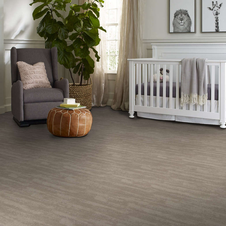 A cozy nursery with a gray armchair, pink pillow, leather ottoman, large plant, white crib with a blanket, and animal art on the walls, all set on a soft, gray floor.