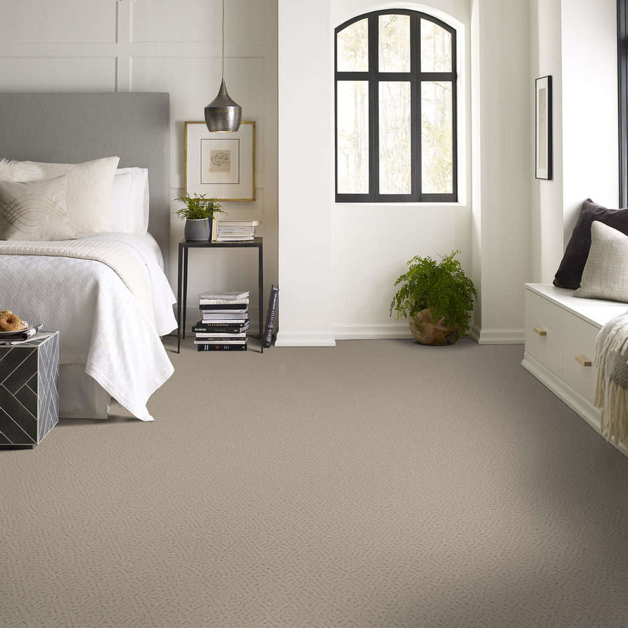 A modern bedroom with a light gray carpet, white bedding, a gray upholstered headboard, black side table, potted plant, window seat, and a large arched window letting in natural light.