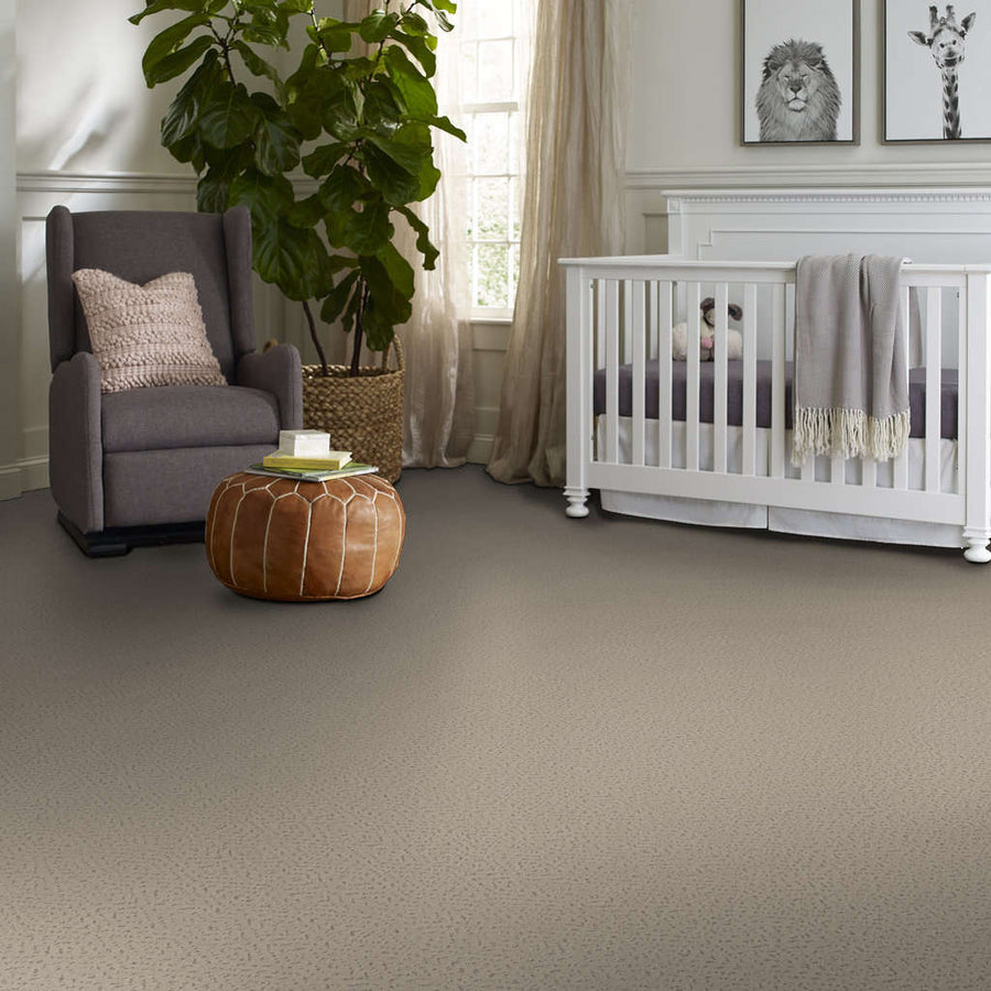 A cozy nursery with a gray armchair, a knitted pillow, a round brown ottoman, a white crib with a gray blanket, a large green plant, and animal art prints on the wall. Sunlight streams in through a window.