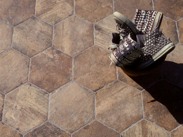 A pair of high-top sneakers with star and studded designs rest in a bowl on a floor of Chicago 9x11 Brick Hex Tile. Sunlight casts shadows, highlighting the texture of both the tiles and the shoes.