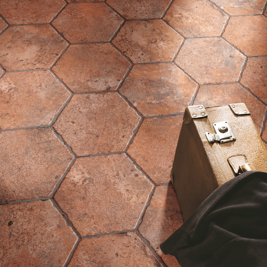 A section of Chicago 9x11 Brick Hex Tile in a reddish-brown shade covers the floor, with sunlight casting soft shadows. A vintage brown suitcase and a dark fabric are partially visible in the bottom right corner.