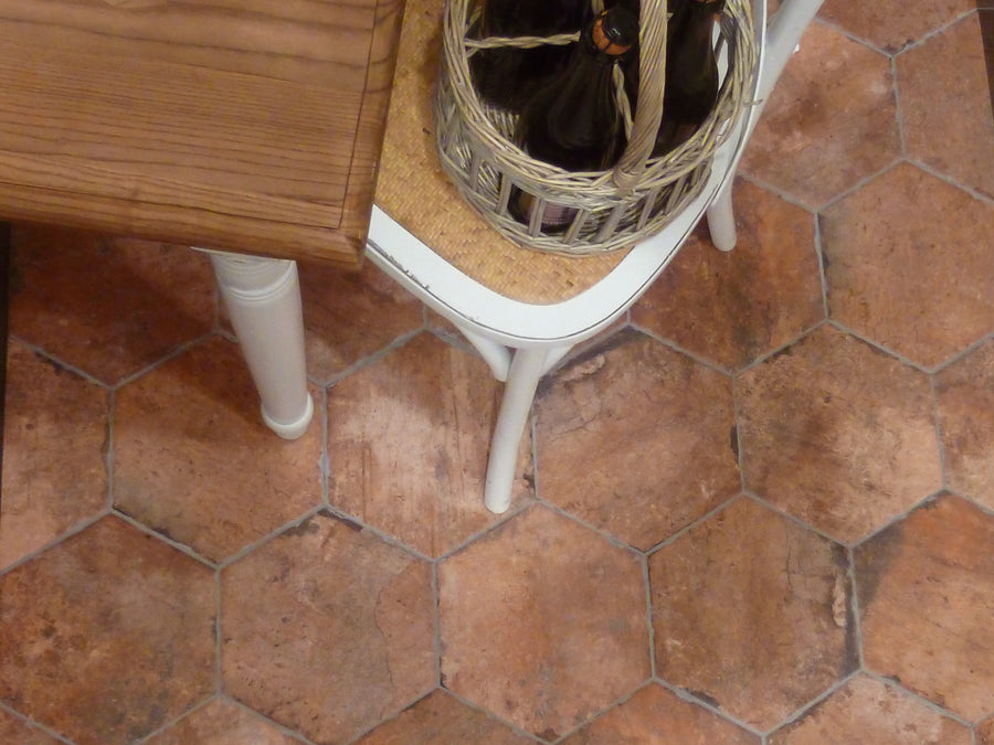 A close-up of a terracotta hexagonal tile floor, with the corner of a wooden table and a white chair holding a wicker basket containing dark glass bottles.
