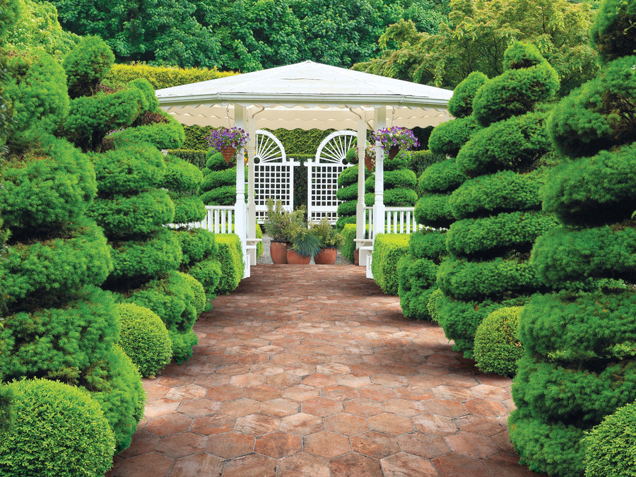 A stone pathway leads through lush, manicured greenery to a white gazebo with a peaked roof. Hanging flower baskets and potted plants decorate the gazebo, surrounded by neatly trimmed bushes and trees.