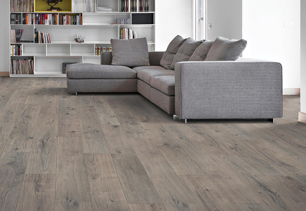 A modern living room with grey wood flooring, a large grey sectional sofa, and a built-in bookshelf filled with books and decor against the back wall. Natural light brightens the space.