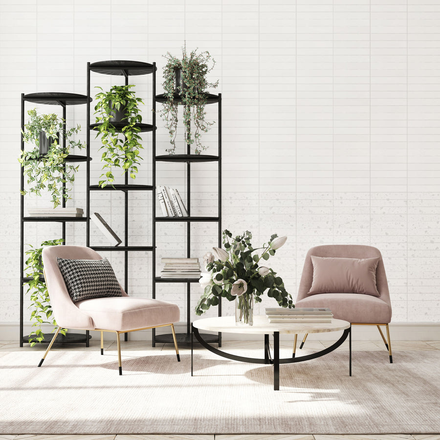Modern living room with two blush pink chairs, a round coffee table with books and a vase of flowers, and a black open shelving unit with green plants and books against a white tiled wall.