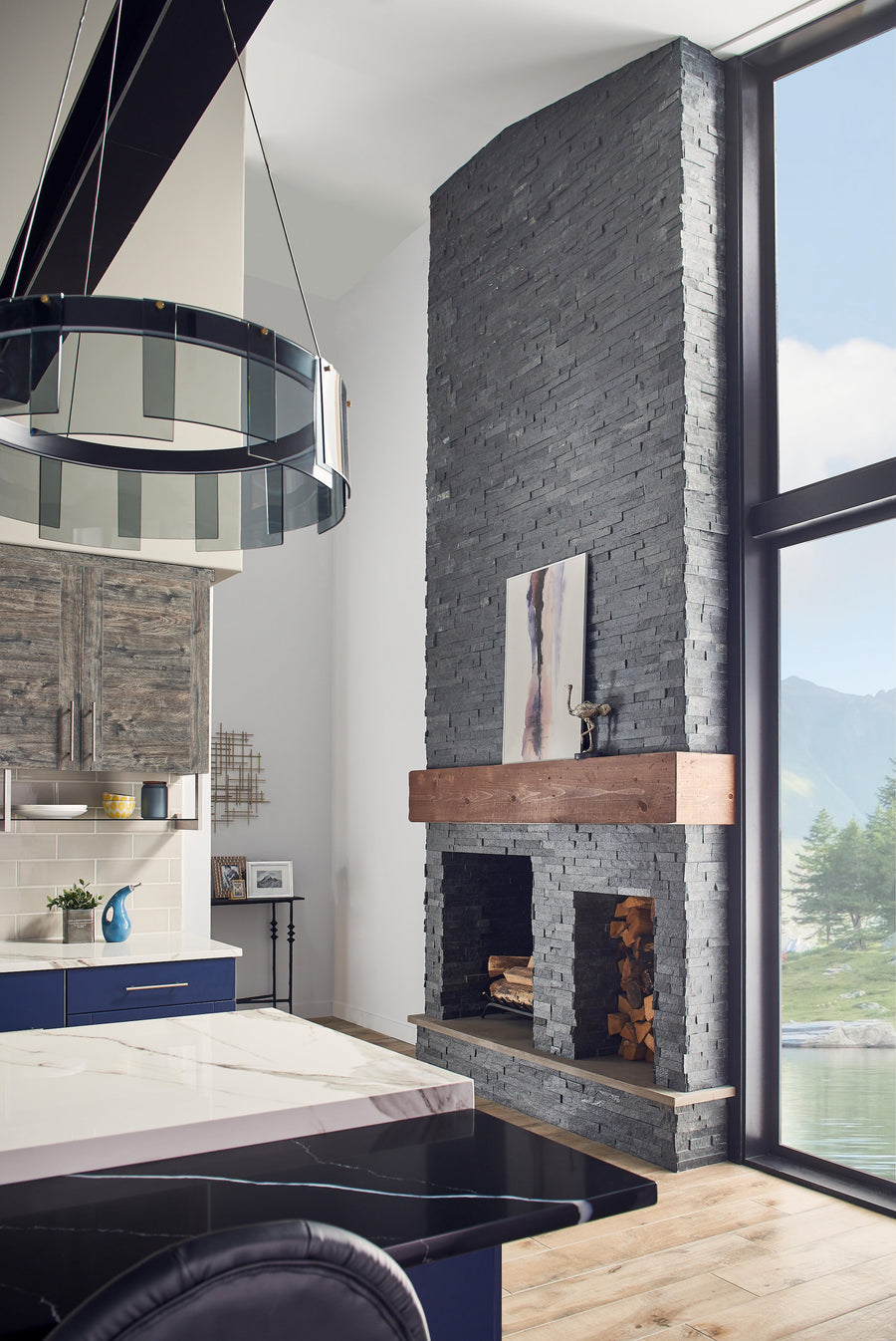 A modern living space with a tall, gray stone fireplace, wooden mantel, and artwork above. Large windows reveal a scenic mountain view. The room features blue cabinets, a marble countertop, and a contemporary chandelier.