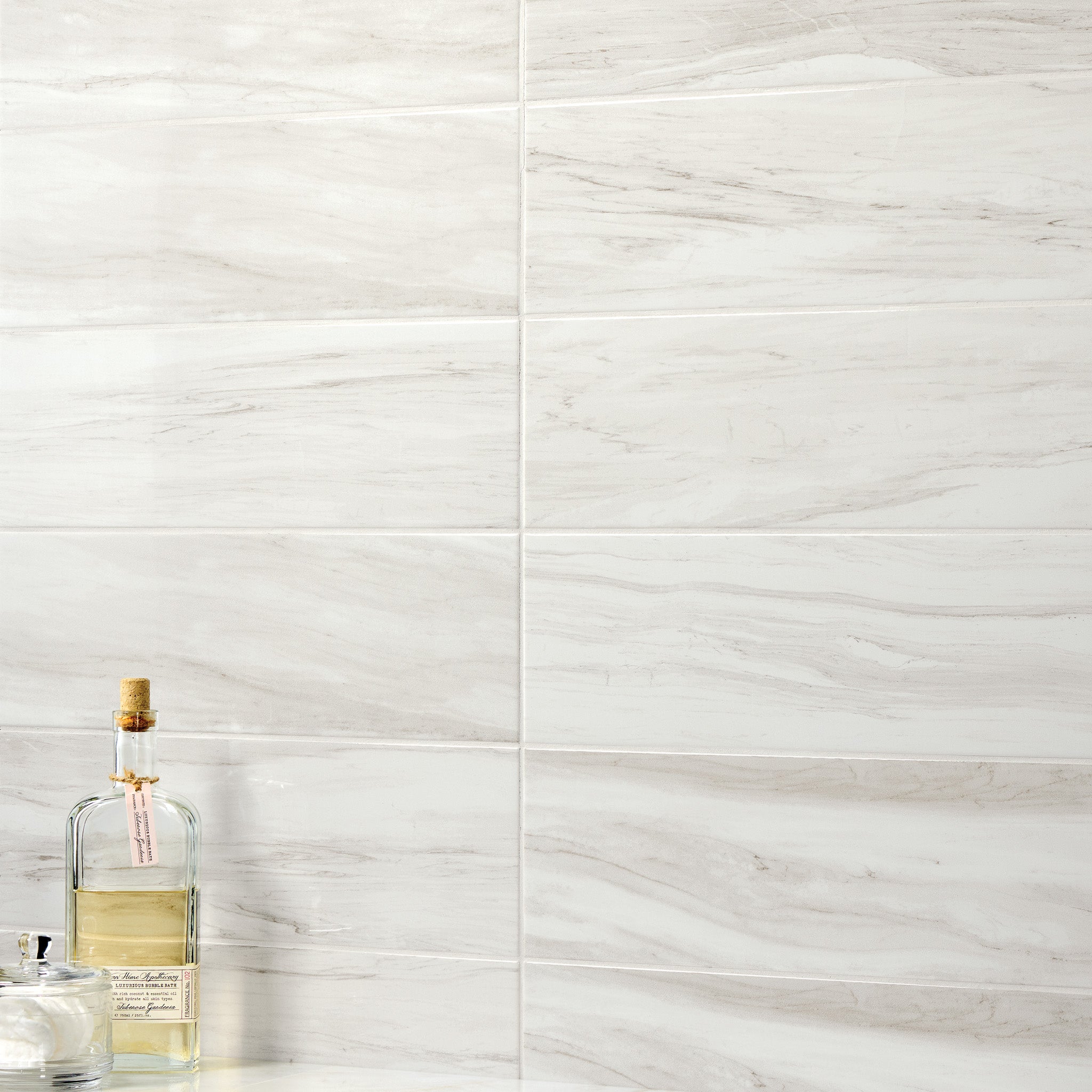 
		                            	Skyline 6x18 Glossy Porcelain Tiles with a white marble aesthetic and grey veining are installed in a horizontal pattern, with a glass bottle and jar sitting on a surface in the foreground.
		                            