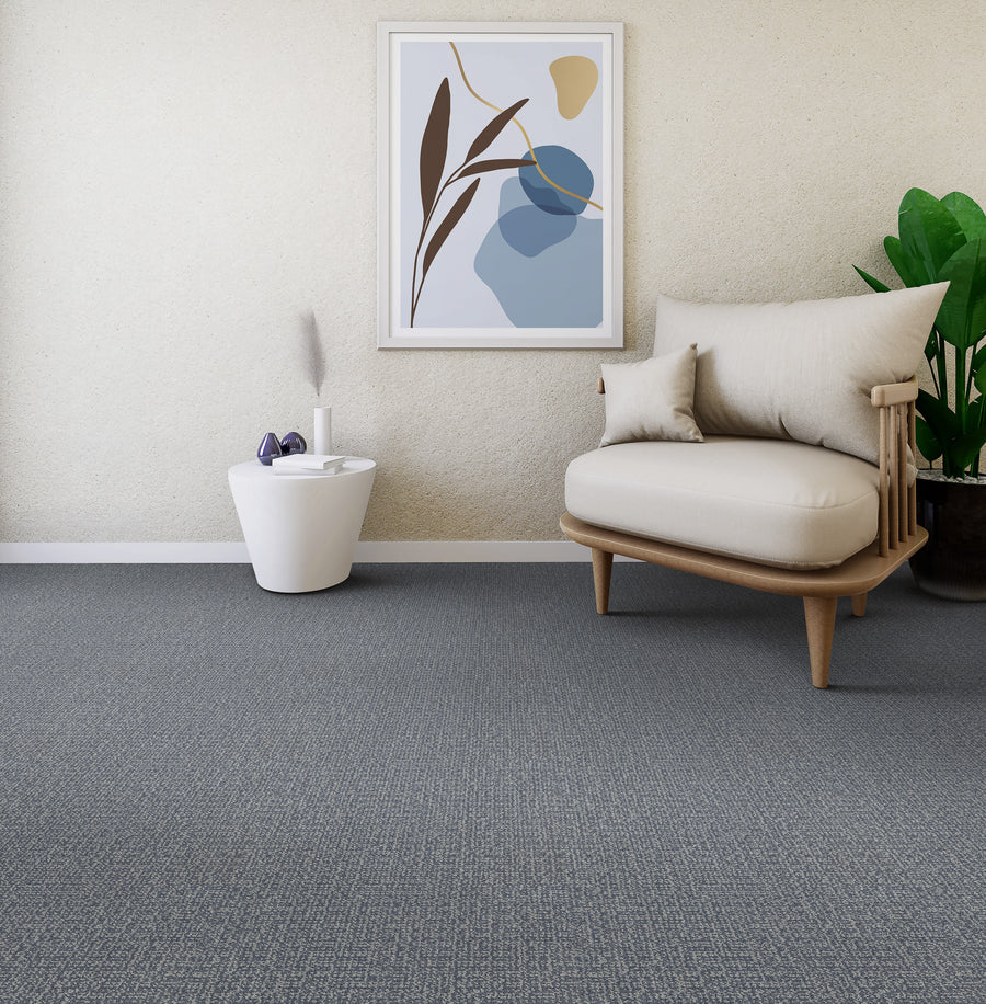 A modern, minimal living space with a beige armchair, a small white side table holding decor, a potted plant, grey carpet, and abstract art on a light-colored wall.