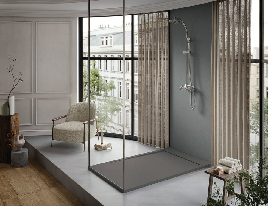 Modern, minimalist bathroom with a low-profile NEOStone Linear End Drain Shower Base in the walk-in shower near large curved windows. Neutral tones, wood floors, a cozy armchair, and soft natural light create a serene retreat with city views.