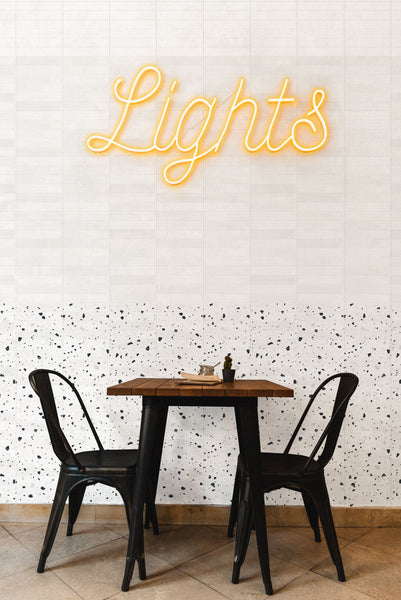 A small wooden table with two black metal chairs sits against a white tiled wall with a terrazzo pattern and a yellow neon sign above that reads Lights.