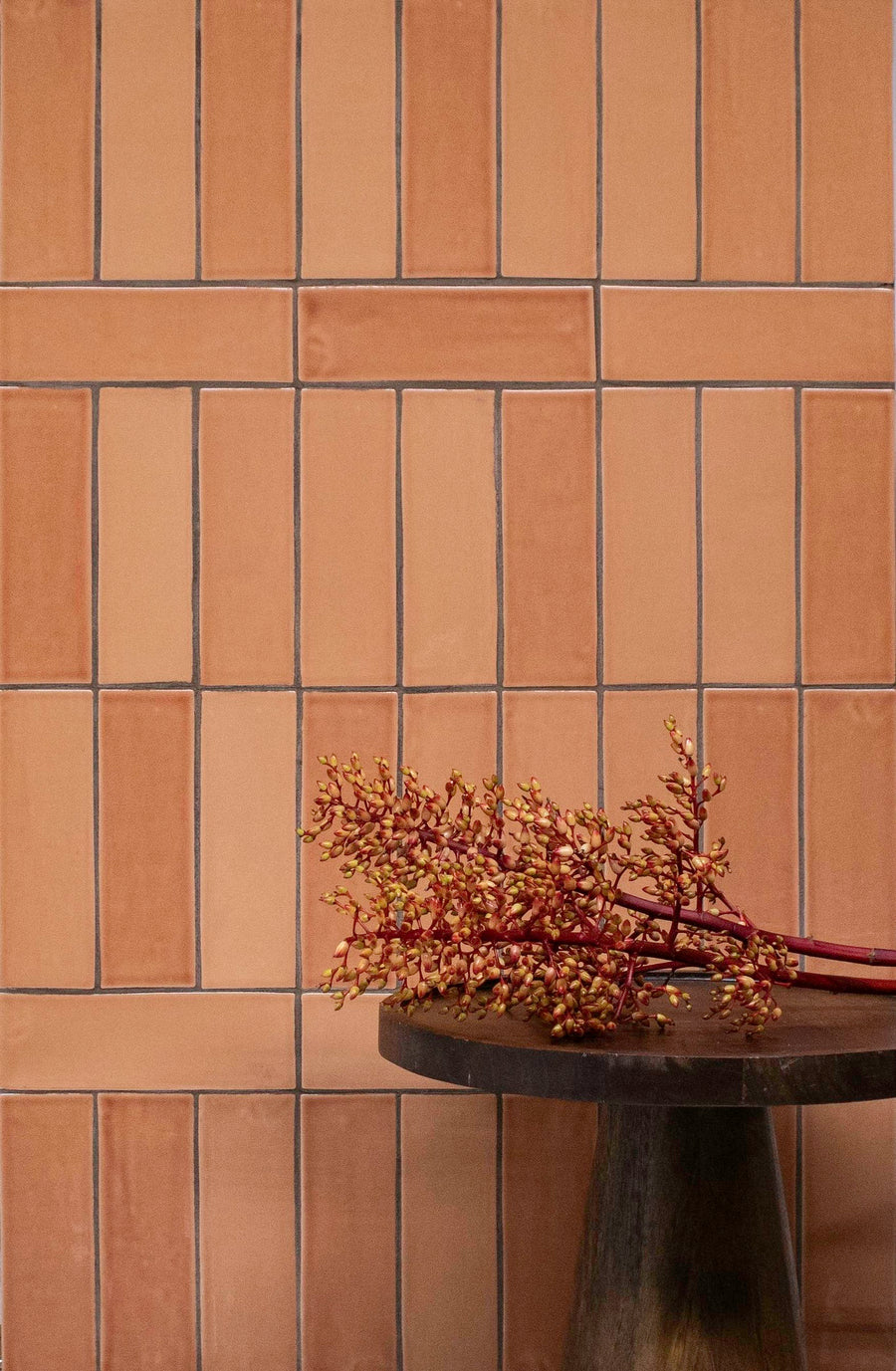 A brown tiled wall with a vertical and horizontal pattern, behind a dark wooden round table holding a branch with small red and yellow berries.