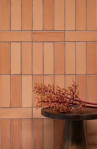 A brown tiled wall with a vertical and horizontal pattern, behind a dark wooden round table holding a branch with small red and yellow berries.
