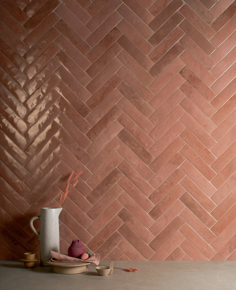 A herringbone pattern wall made of glossy and matte pink tiles, with a beige surface below holding a white ceramic pitcher, a napkin, and small tableware.