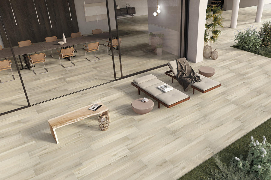 Modern outdoor patio with light wood-look tile flooring, lounge chairs with cushions, a small round side table, a wooden bench with a book, and greenery along the edges, next to a glass-walled dining area.