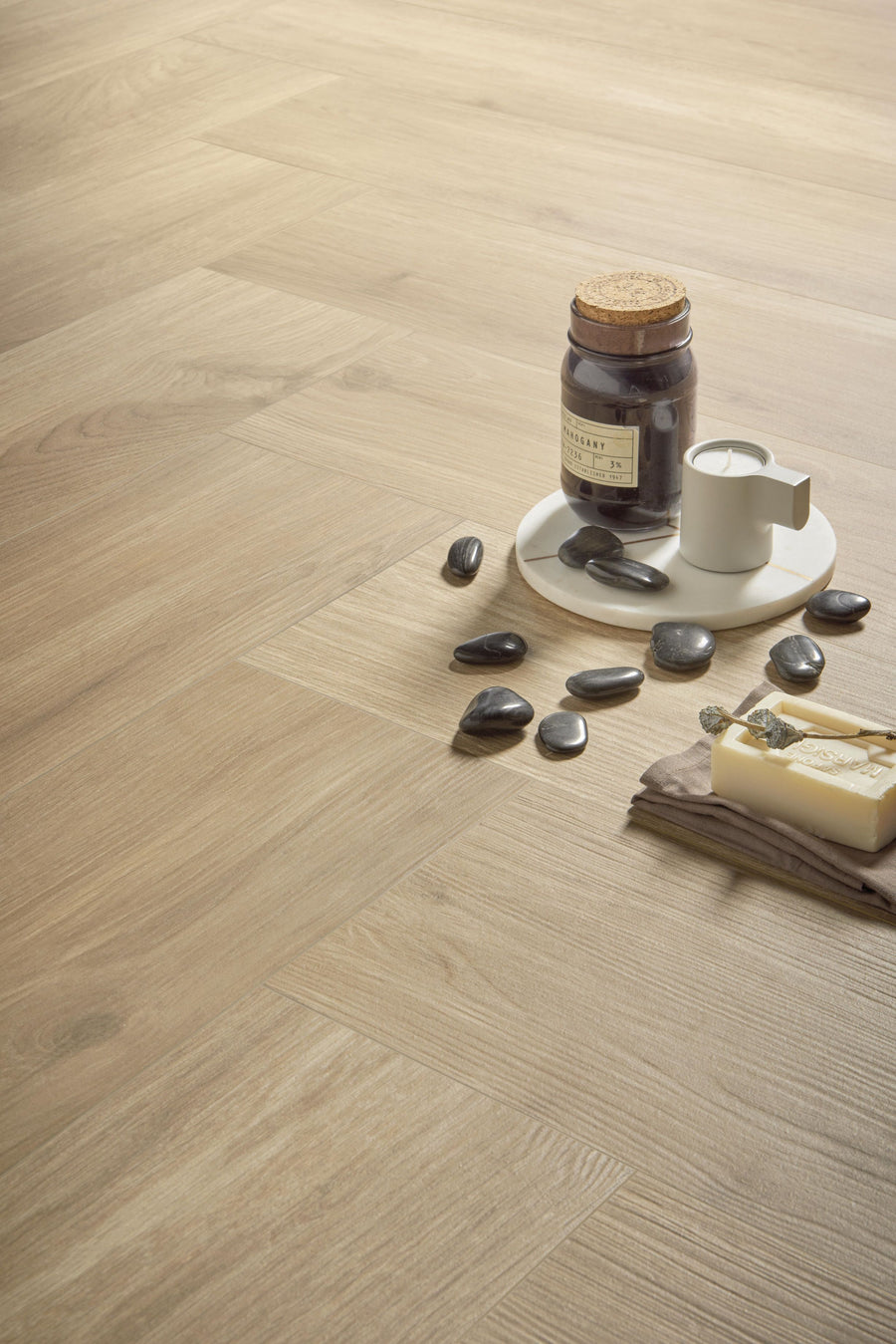 A light wood floor with a white tray holding a jar, a candle, black decorative stones, and a bar of soap on a folded cloth. The arrangement creates a calm, spa-like atmosphere.