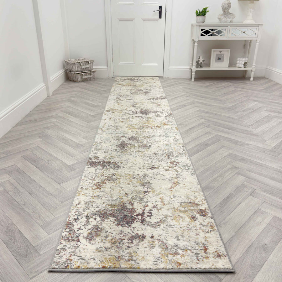 The Couture 52023-6616 Runner Rug, featuring a mottled abstract pattern in neutral tones, is placed on a light herringbone wood floor in a bright hallway with white walls, a white door, and a small white console table.